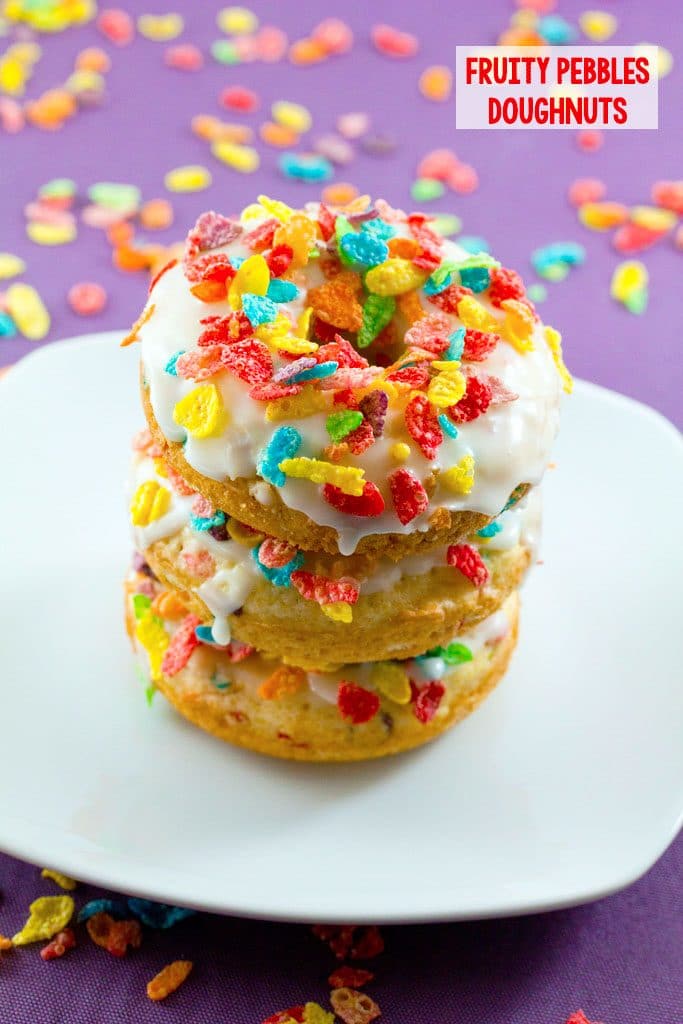 Three Fruity Pebbles Doughnuts stacked on a white plate with Fruity Pebbles scattered on a purple background