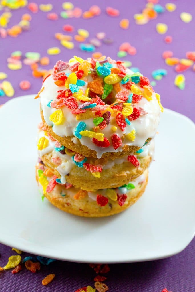 Three Fruity Pebbles Doughnuts stacked on a white plate with a purple background with Fruity Pebbles sprinkled around