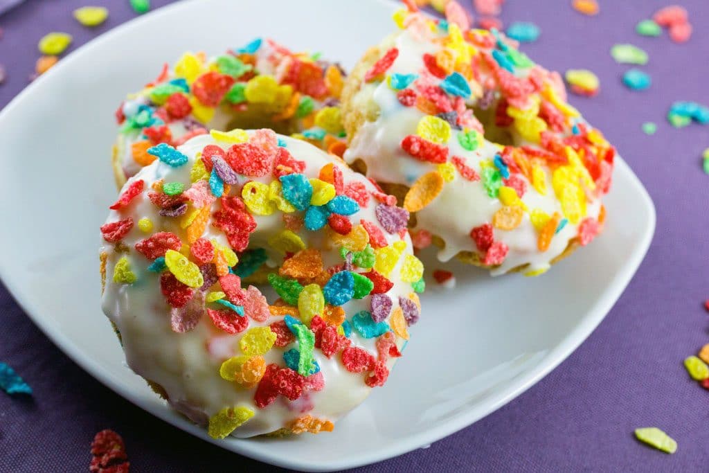 Three Fruit Pebbles Doughnuts laying on a white plate with a purple background and Fruity Pebbles sprinkled around