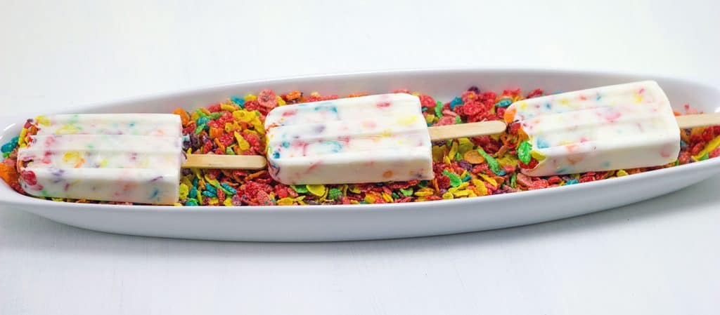 Horizontal image featuring three Fruity Pebbles Greek yogurt popsicles on a white plate in a bed of Fruity Pebbles