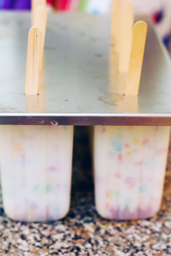 Popsicle molds filled with Greek yogurt, milk, and Fruity Pebbles mixture upside down with popsicle sticks in