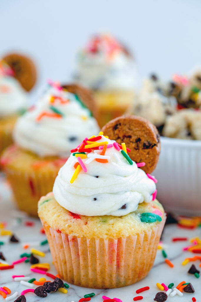 Head-on view of a funfetti cookie dough cupcake with chocolate chip frosting and a mini chocolate chip cookie on top, surrounded by sprinkles and a bowl of cookie dough.