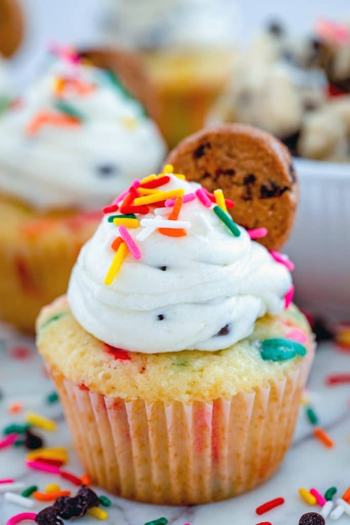 Head-on close-up view of funfetti cookie dough cupcake topped with frosting, sprinkles, and mini chocolate chip cookie with sprinkles and chocolate chips, more cupcakes, and bowl of cookie dough in the background