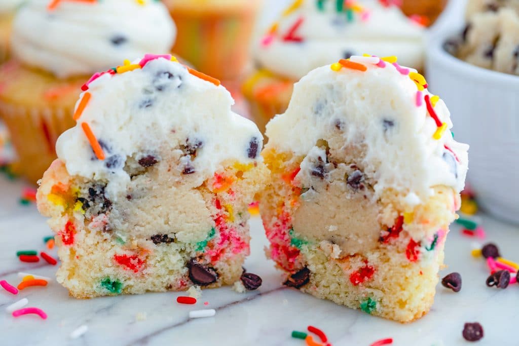 Landscape view of a funfetti cookie dough cupcake cut in half with cookie dough center revealed with sprinkles, mini chocolate chips and more cupcakes in background