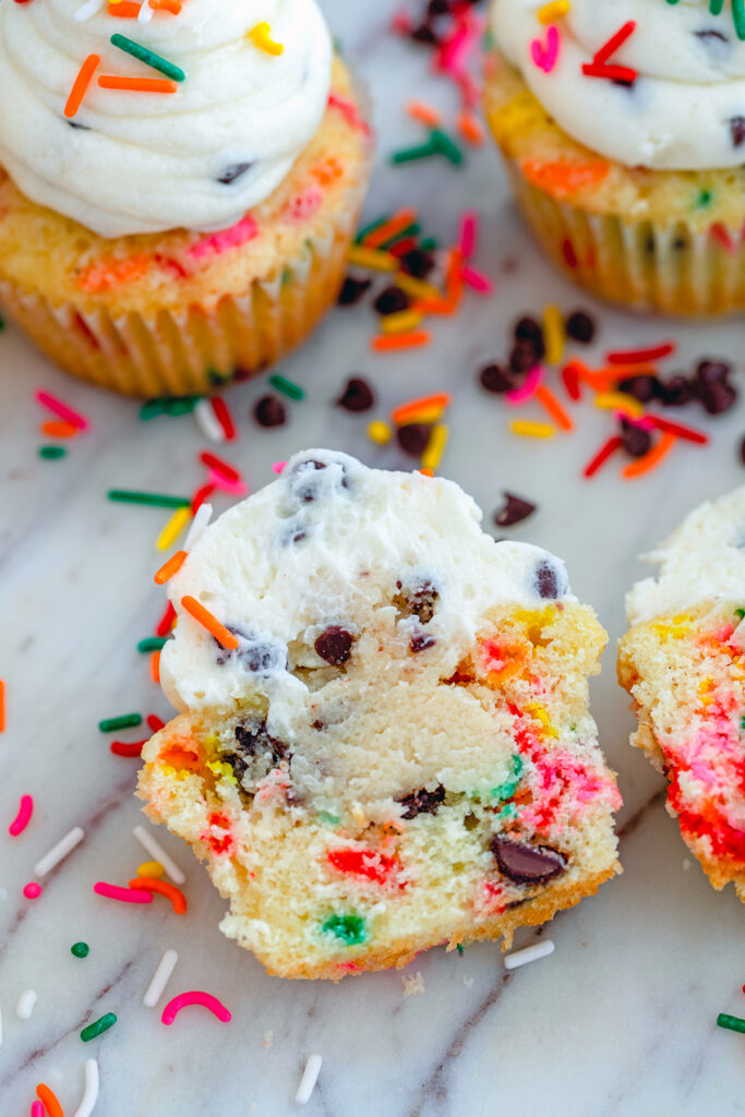 Overhead view of funfetti cookie dough cupcake cut in half with cookie dough center showing, surrounded by sprinkles, chocolate chips, and more cupcakes in background.