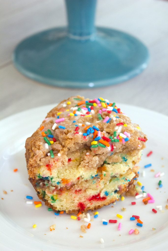 Overhead view of slice of funfetti coffee cake topping with crumb topping and rainbow sprinkles on a white plate with sprinkles scattered around