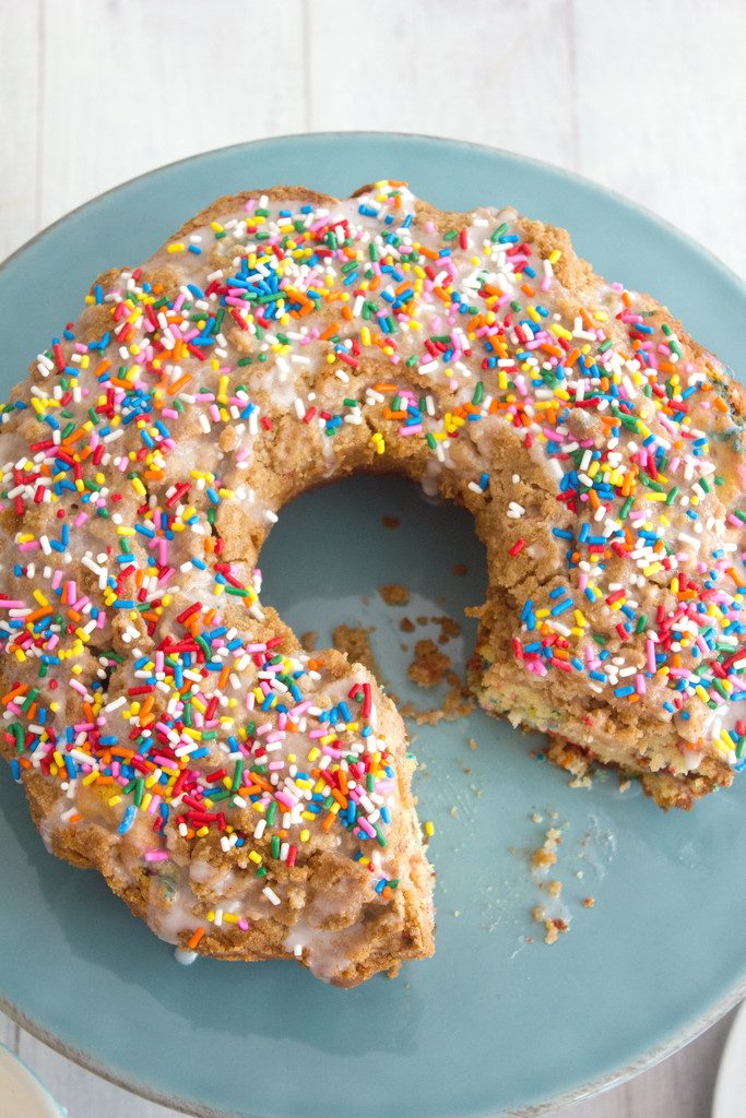 Overhead view of funfetti coffee cake topped with icing and rainbow sprinkles with a slice removed on a turquoise cake platter