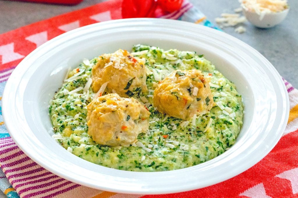 Landscape view of white bowl filled with kale polenta topped with three garlic habanero chicken meatballs on a colorful placemat with tablespoon of parmesan cheese in the background