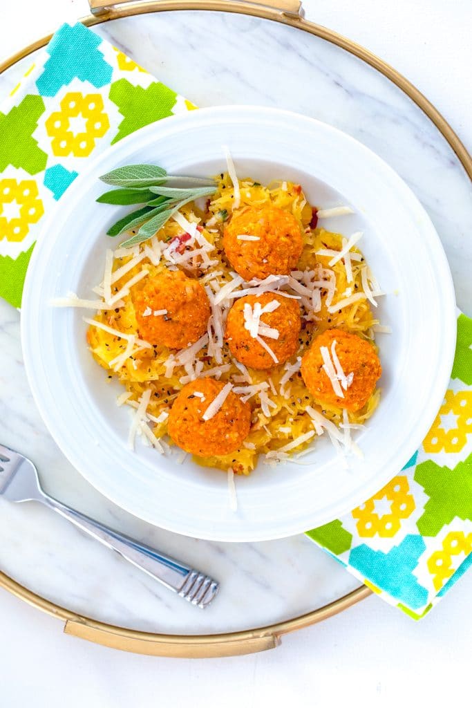 Bird's eye view of white bowl of garlicky spaghetti squash with pumpkin chicken meatballs with parmesan and sage on a marble surface with colorful napkin and fork