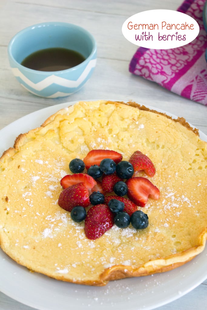 Head-on view of a large German pancake topped with berries and powdered sugar with a bowl of maple syrup in the background and recipe title at top