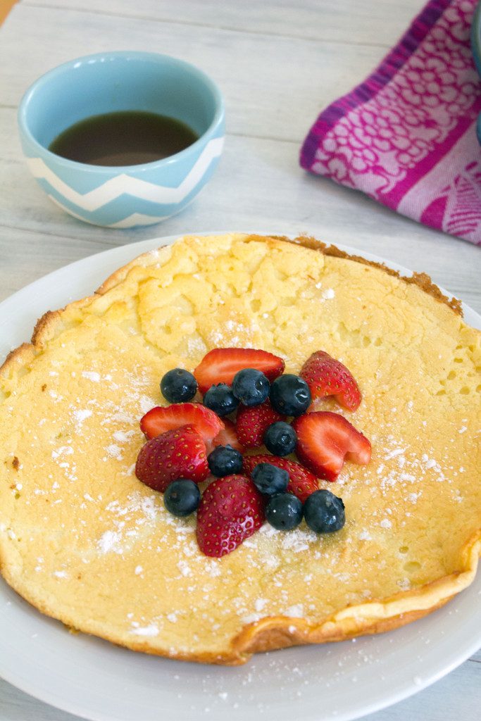 Head-on view of a large German pancake topped with berries and powdered sugar with a bowl of maple syrup in the background