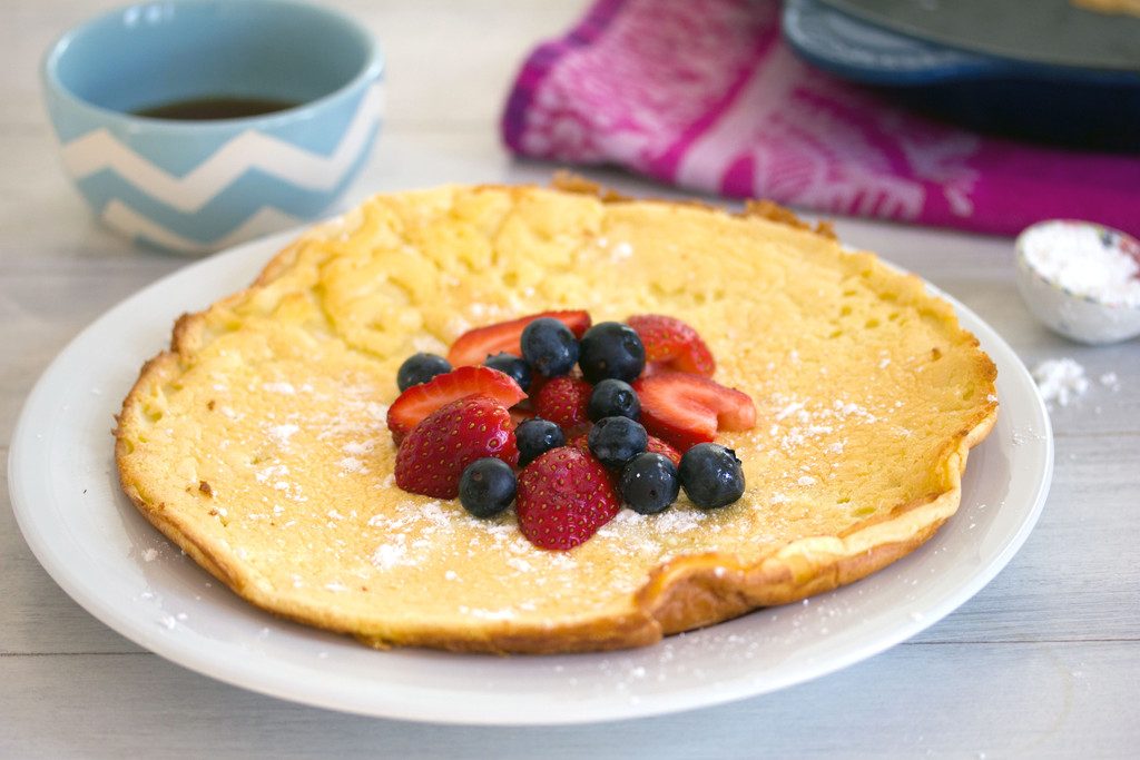 Head-on landscape view of a large German pancake topped with berries and powdered sugar with bowl of maple syrup, skillet, and ceramic tablespoon in the background