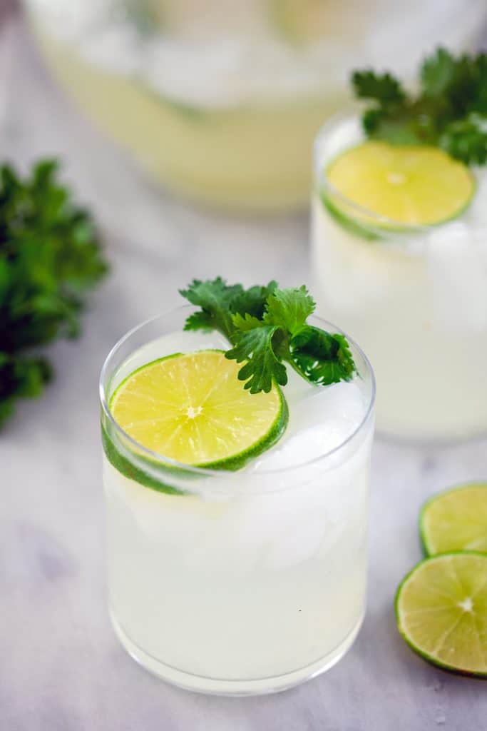 Head-on view of gin lime rickey in a rocks glass with lime round and cilantro, a second cocktail in the background, along with additional lime wedges, cilantro, and a pitcher