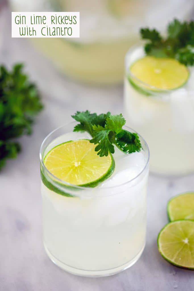 Head-on view of gin lime rickey in a rocks glass with lime round and cilantro, a second cocktail in the background, along with additional lime wedges, cilantro, and a pitcher with recipe title at the top of the image