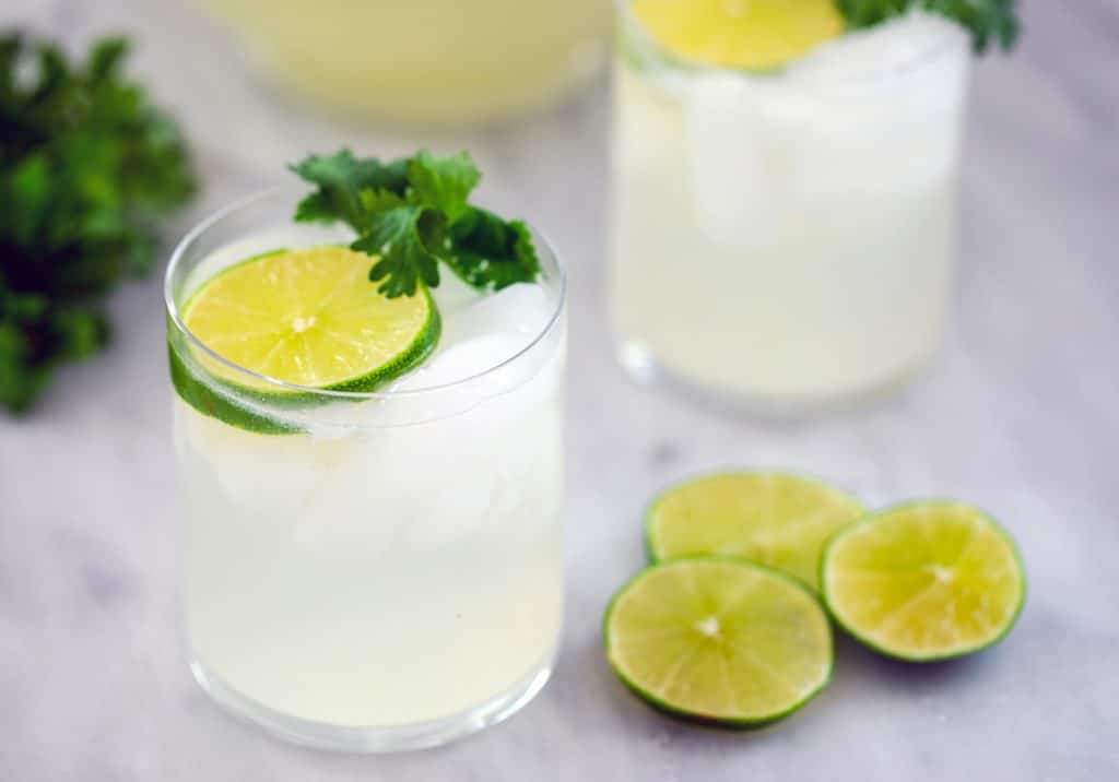 Landscape view of two gin lime rickeys with lime rounds and cilantro with additional lime rounds and a bunch of cilantro in the background