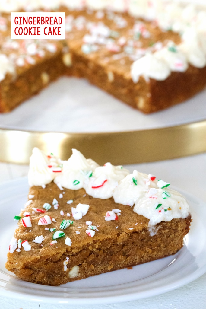 Head-on view of a slice of gingerbread cookie cake with frosting and peppermint crumbles on a white plate with full cookie cake in the background with recipe title at top