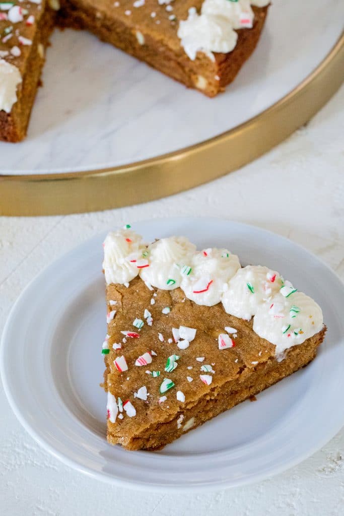 Overhead view of slice of gingerbread cookie cake on a white plate with buttercream frosting and red and green crushed candy canes 