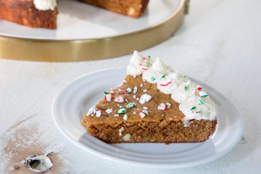 Landscape view of slice of gingerbread cookie cake on a white plate with buttercream frosting and crushed red and green candy canes