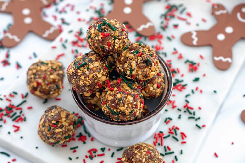 Landscape overhead view of gingerbread energy balls in a small bowl with more energy bites, red and green sprinkles, and gingerbread garland in background