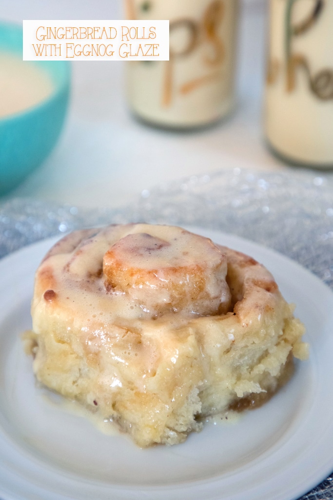 Head-on view of a gingerbread roll with eggnog icing on a small plate with glasses of eggnog and bowl of eggnog glaze in the background and recipe title at top