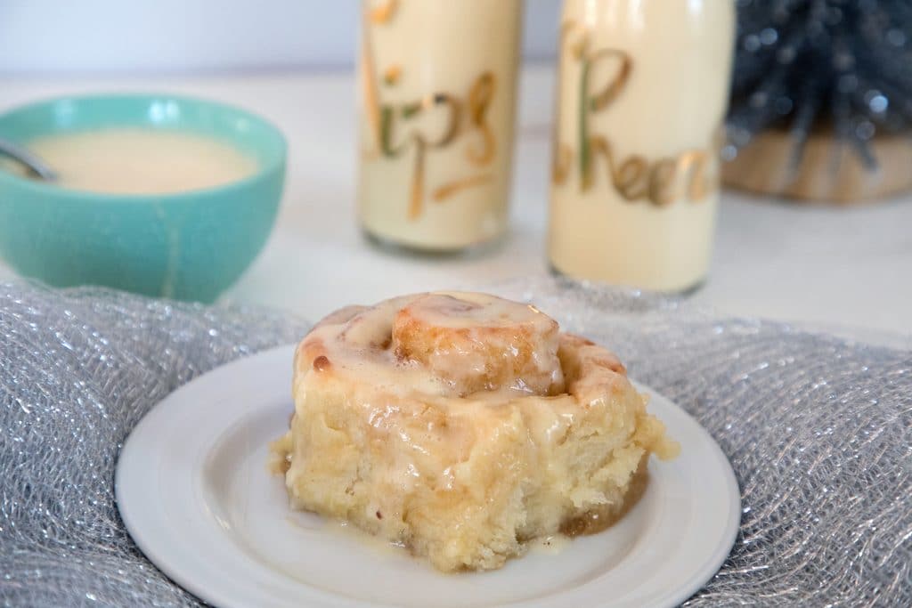 Landscape head-on view of a gingerbread roll with eggnog glaze on a white plate with silver tinsel with glasses of eggnog and bowl of eggnog glaze in background