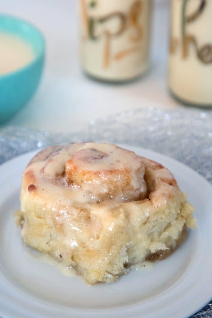 Head-on view of a gingerbread roll with eggnog icing on a small plate with glasses of eggnog and bowl of eggnog glaze in the background