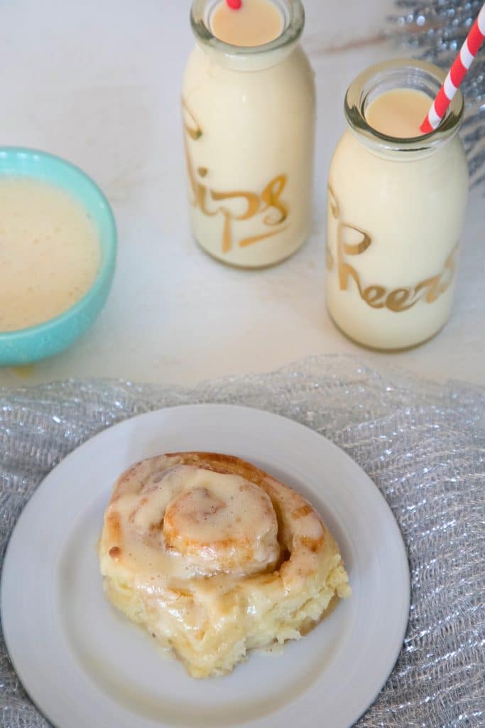 Overhead view of a gingerbread eggnog roll on a small white plate with glasses of eggnog and bowl of eggnog glaze in the background