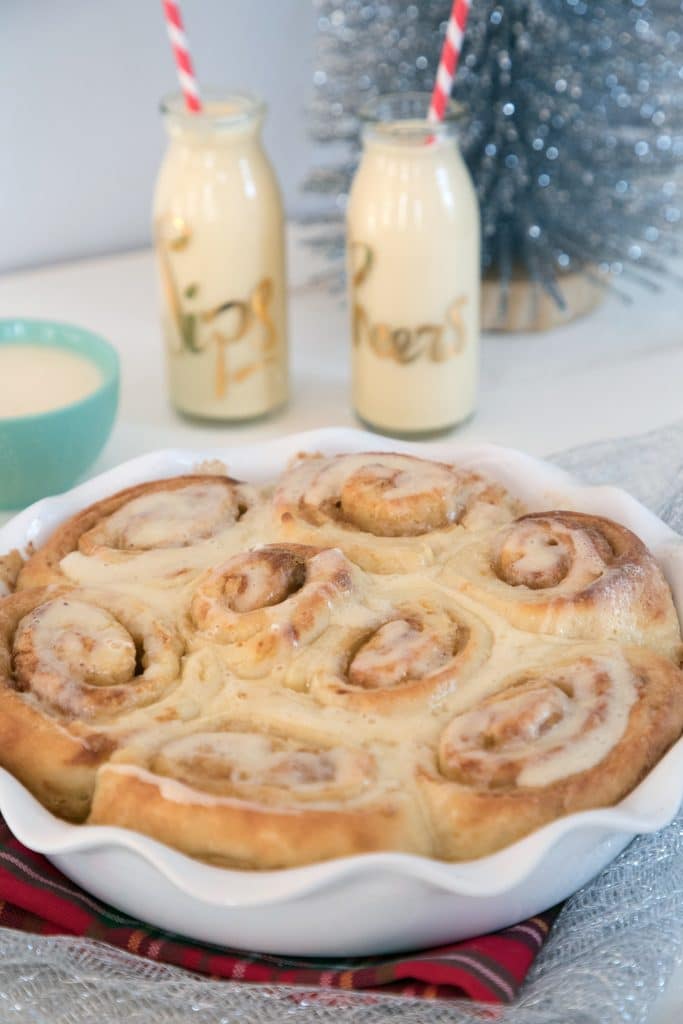 Head-on view of a pie plate of gingerbread rolls with eggnog glaze with glasses of eggnog, bowl of eggnog glaze, and silver mini tree in background