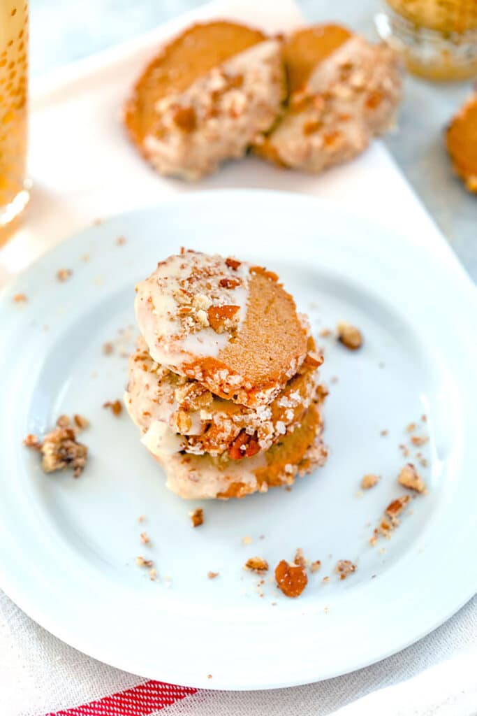 Head-on view of a stack of three gingerbread shortbread cookies with eggnog glaze and pecans on a white plate with pecan crumbles and more cookies in the background