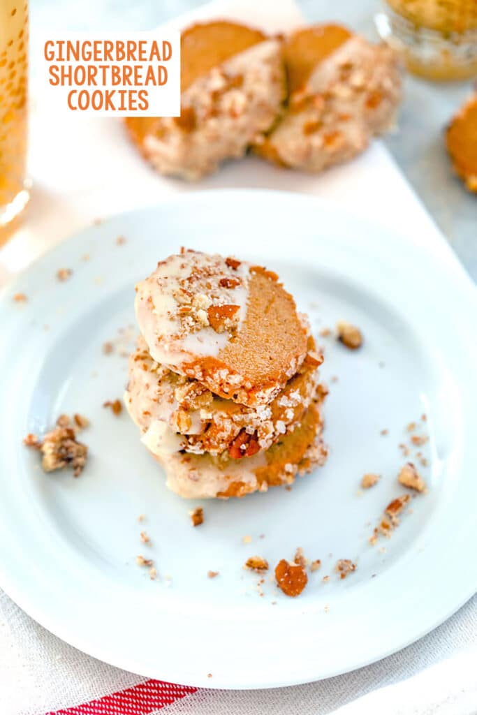 Head-on view of a stack of three gingerbread shortbread cookies with eggnog glaze and pecans on a white plate with pecan crumbles, cookies in the background and recipe title at the top