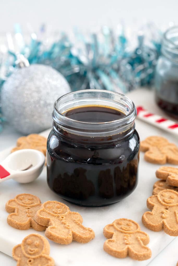 Small jar of gingerbread syrup on a marble surface with gingerbread men and ornaments all around.