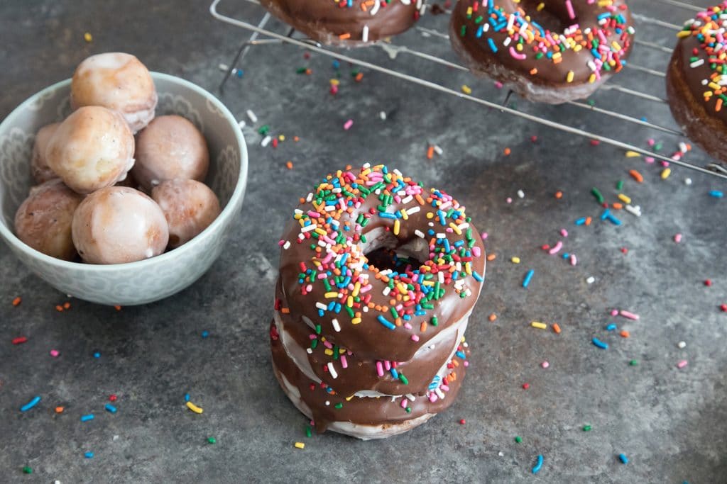 Landscape view of stack of glazed coffee chocolate frosted donuts with sprinkles with baking rack with more donuts in the background and bowl of donut holes