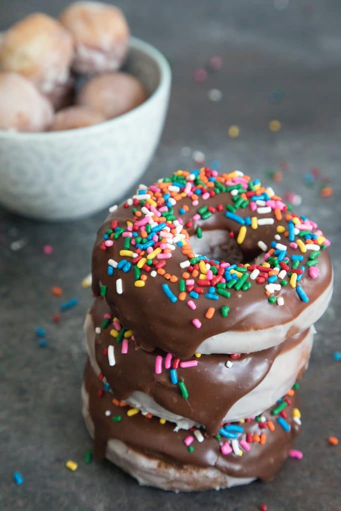 Close-up head-on view of stack of three glazed coffee chocolate frosted donuts with sprinkles with bowl of donut holes in the background