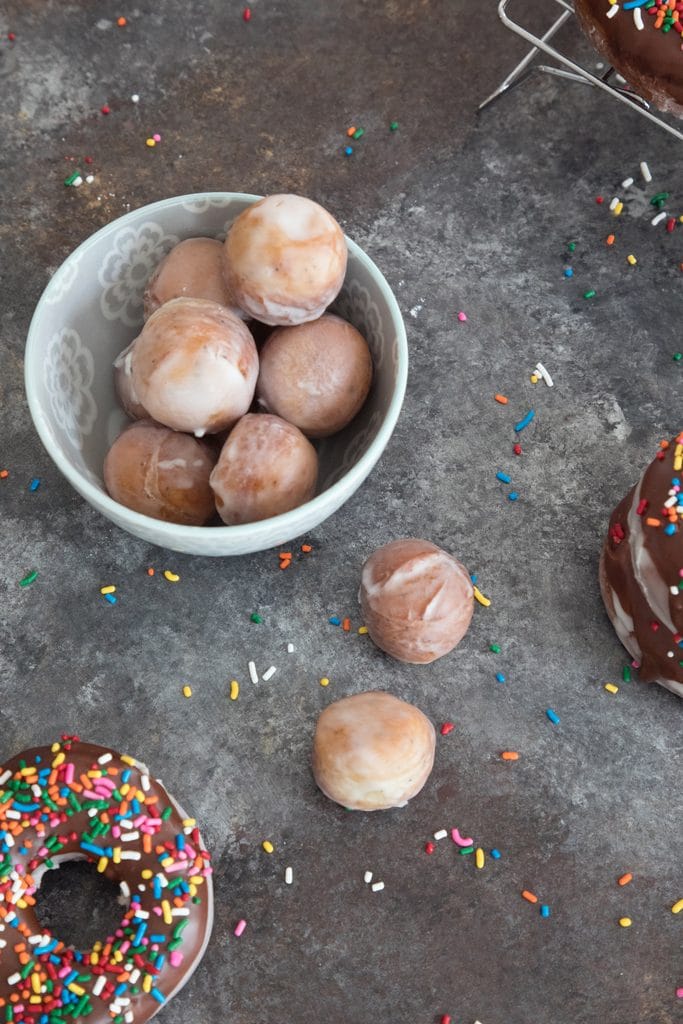 Bird's eye view of bowl filled with glazed coffee donut holes with chocolate frosted donuts with sprinkles in the background