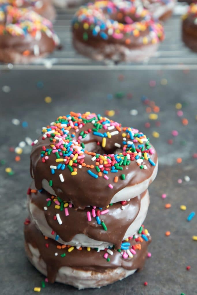 Head-on view of a stack of three glazed coffee chocolate frosted donuts with sprinkles with a baking rack with more donuts in the background