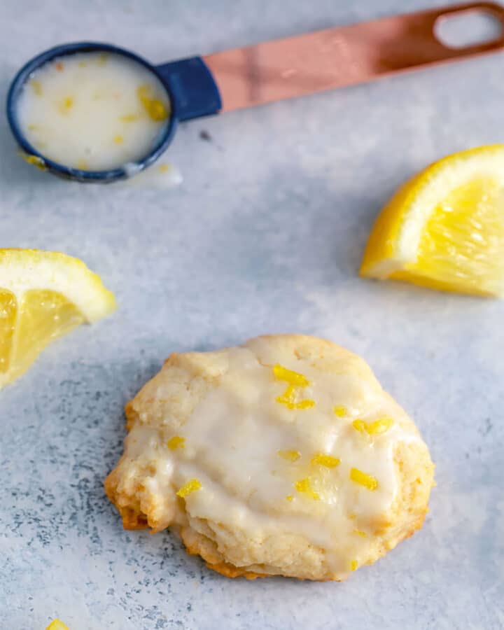 Glazed lemon cookie surrounded by lemon wedges with spoon of lemon glaze in background