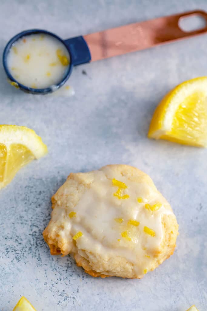 Glazed lemon cookie on a grey surface surrounded by lemon wedges and teaspoon filled with lemon glaze