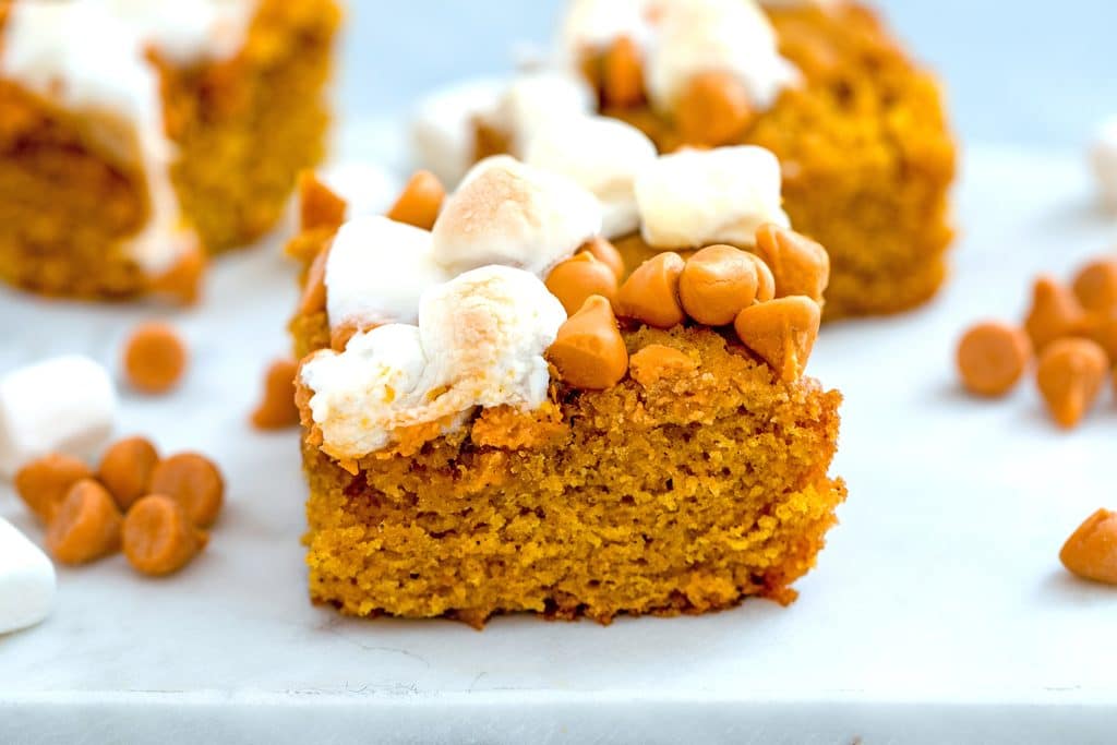 Landscape image featuring close-up view of a pumpkin butterscotch blondie on a white surface with mini marshmallows and butterscotch chips scattered around