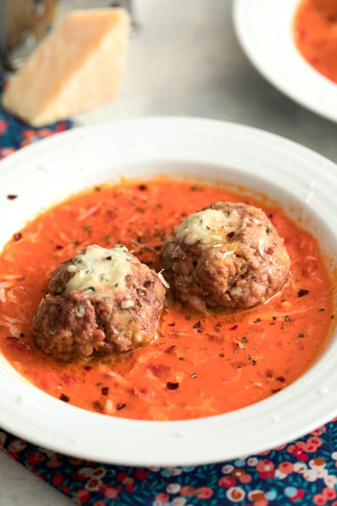 Head-on view of two gorgonzola meatballs in a creamy tomato sauce in a white bowl with second bowl and wedge of parmesan in the back