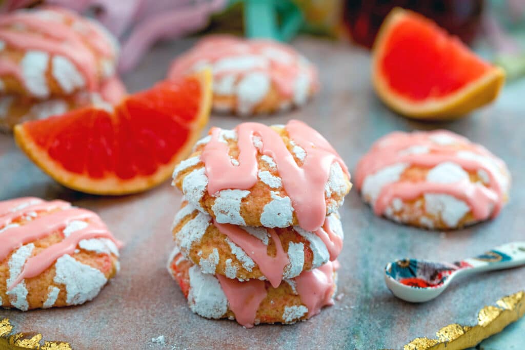 Landscape view of three stacked grapefruit crinkle cookies with more cookies, grapefruit wedges, and spoon of Campari 