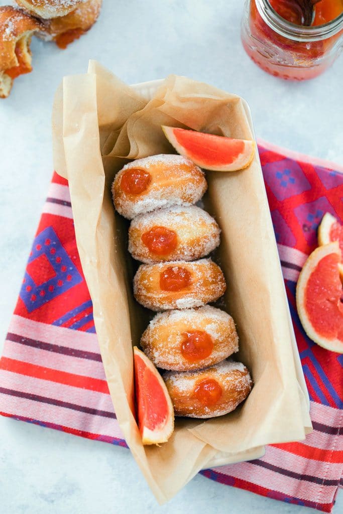 Bird's-eye view of five sugared donuts filled with grapefruit jam in a parchment-lined loaf pan with grapefruit sections and a jar of grapefruit jam