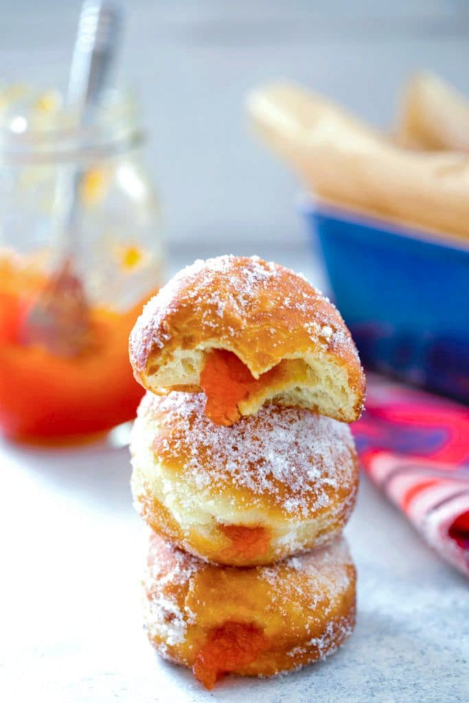 Three sugared donuts filled with grapefruit jam stacked on each other with jelly oozing out and a jar of grapefruit jam in the background