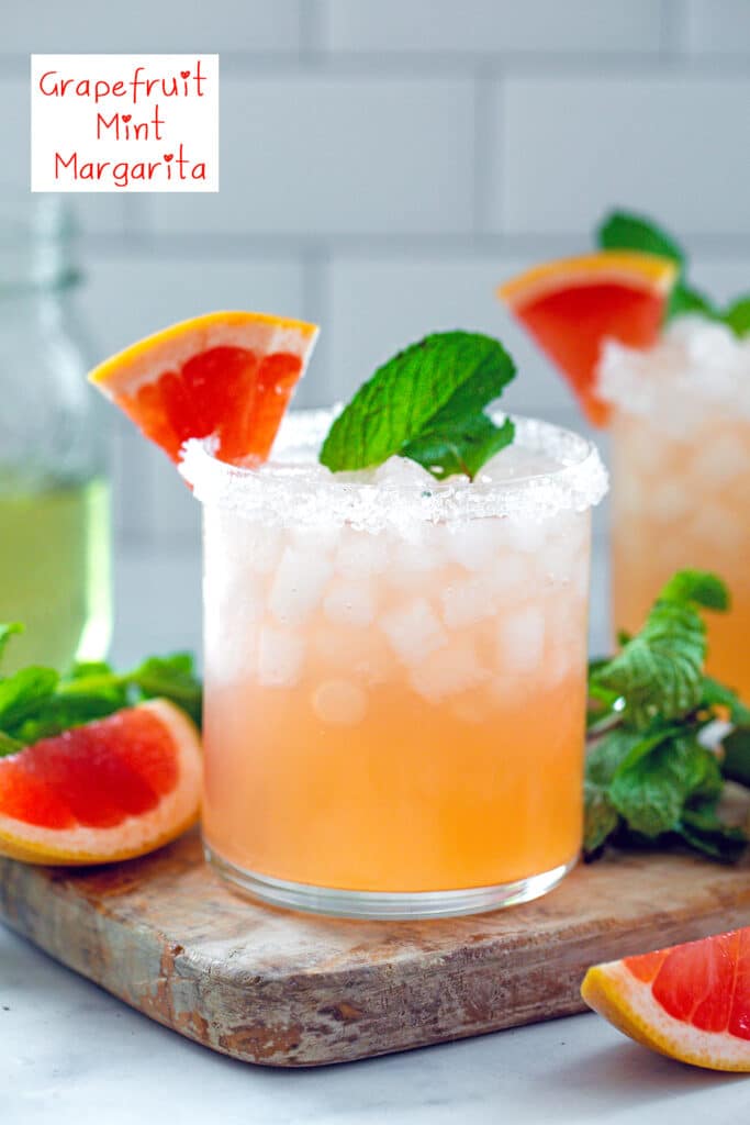 Head-on view of a grapefruit mint margarita with grapefruit slices all around and jar of mint simple syrup in the background and recipe title at top
