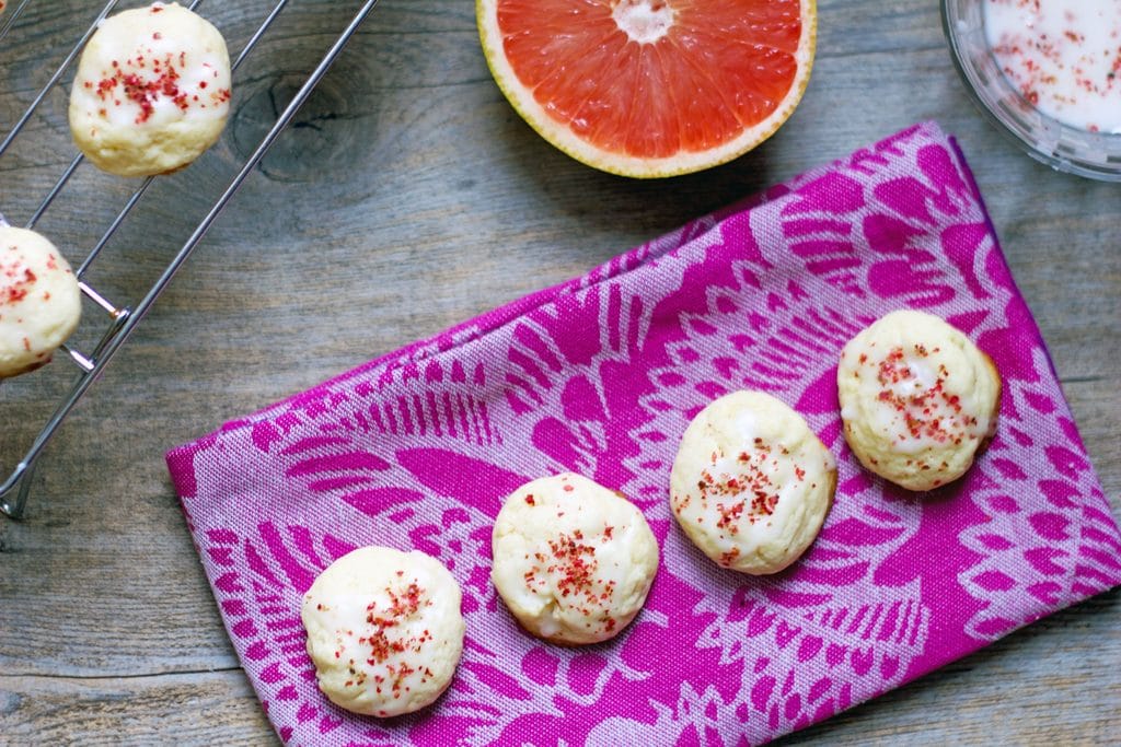 A landscape photo of four grapefruit pink peppercorn cookies lined up on a pink towel with rack with more cookies, icing, and half a grapefruit in the background