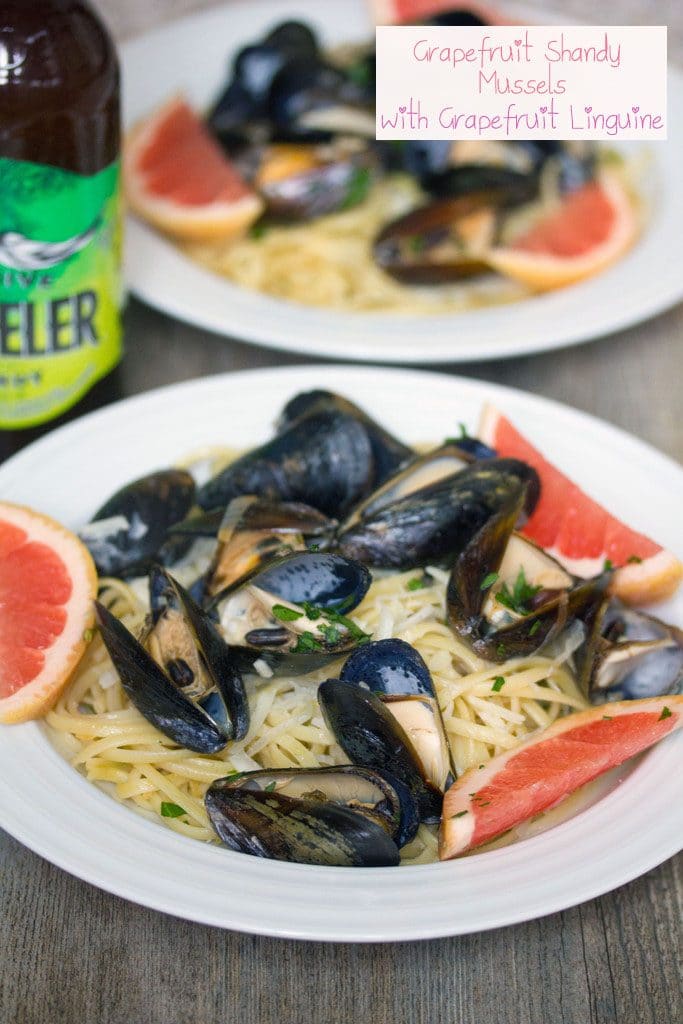 Head-on view of a white bowl of grapefruit shandy mussels over linguine with grapefruit slice garnishes, second bowl in the background, bottle of beer, and recipe title at top