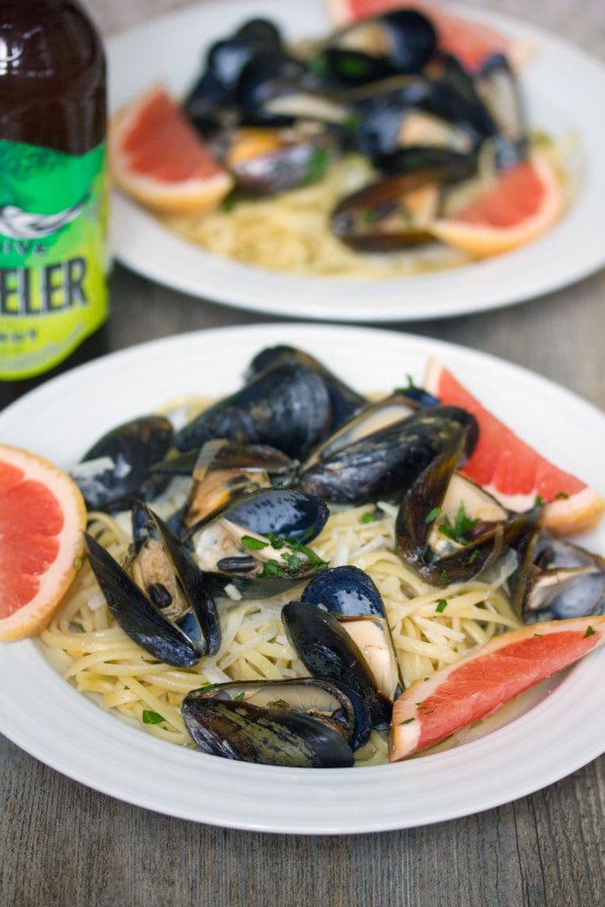 Head-on view of a white bowl of grapefruit shandy mussels over linguine with grapefruit slice garnishes, second bowl in the background, and bottle of beer