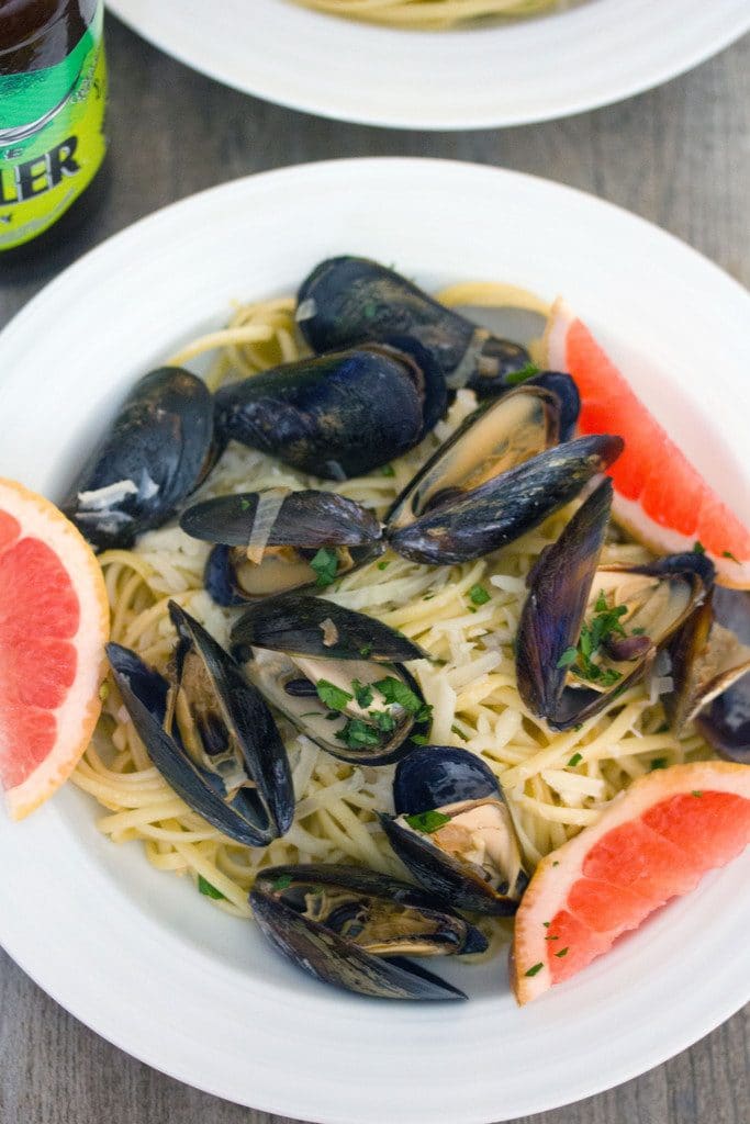 Overhead view of white bowl of grapefruit shandy mussels over linguine with grapefruit wedge garnish and bottle of beer in the background