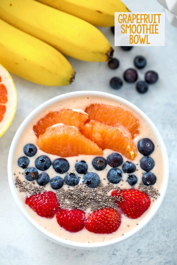 Overhead view of grapefruit smoothie bowl topped with grapefruit sections, blueberries, chia seeds, and sliced strawberries with bananas, blueberries, and grapefruit half in the background and "Grapefruit Smoothie Bowl" text at the top