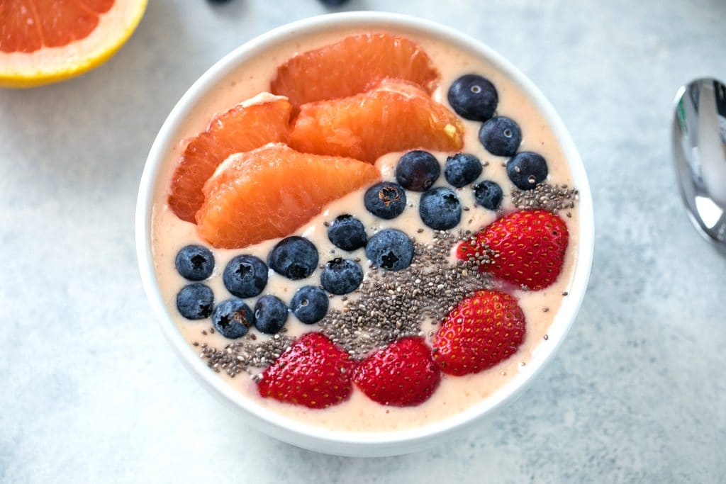 Landscape photo closeup of grapefruit smoothie bowl topped with grapefruit sections, blueberries, chia seeds, and strawberries with spoon in the background