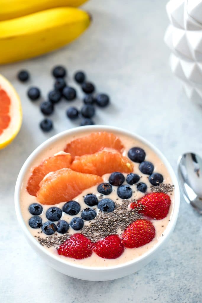 Bird's eye view of grapefruit smoothie bowl topped with grapefruit sections, blueberries, chia seeds, and strawberries with grapefruit half, bananas, blueberries, and spoon in the background